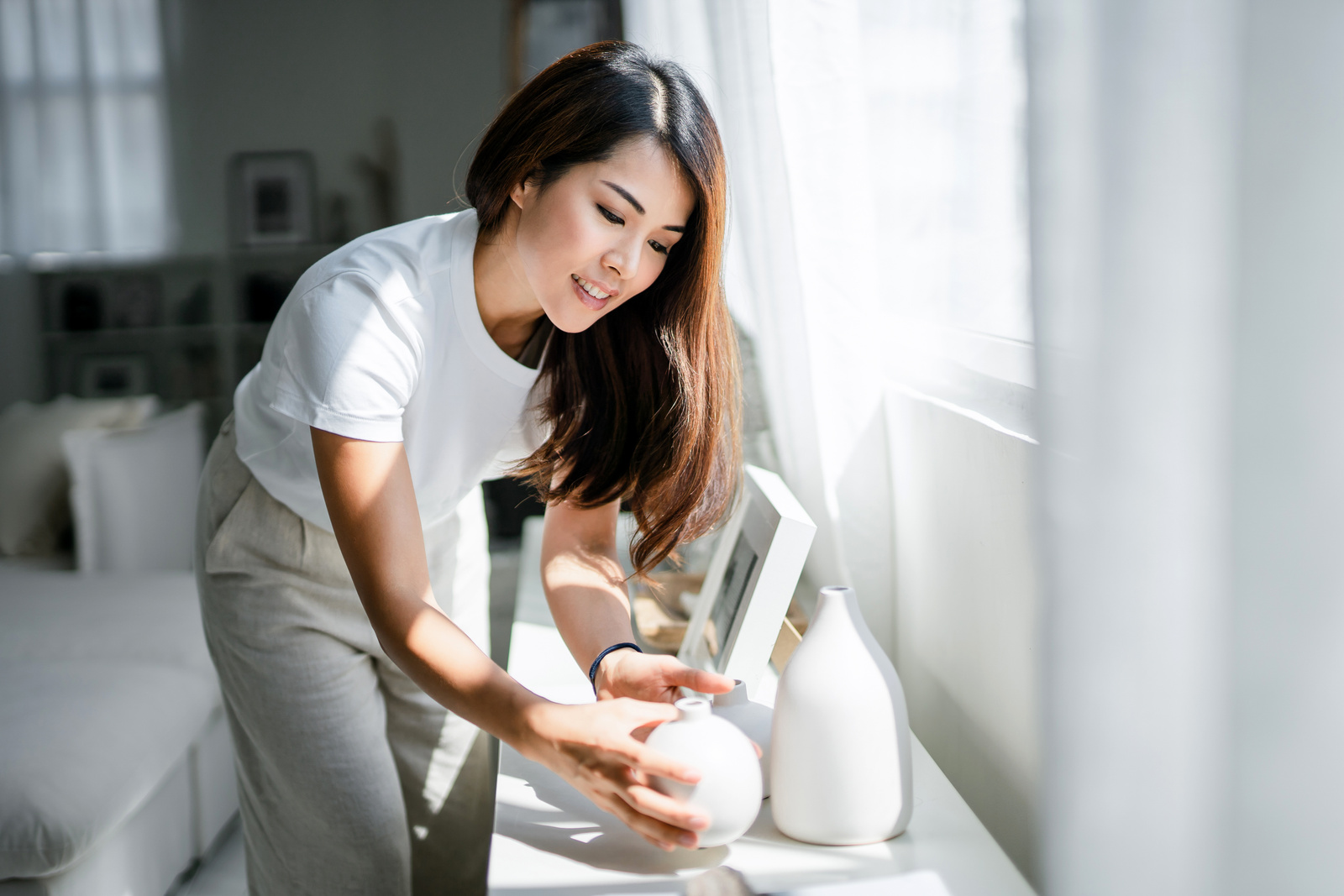 Young Asian woman enjoys her time at home, decorating and organising picture frames and vases on the shelf by the window